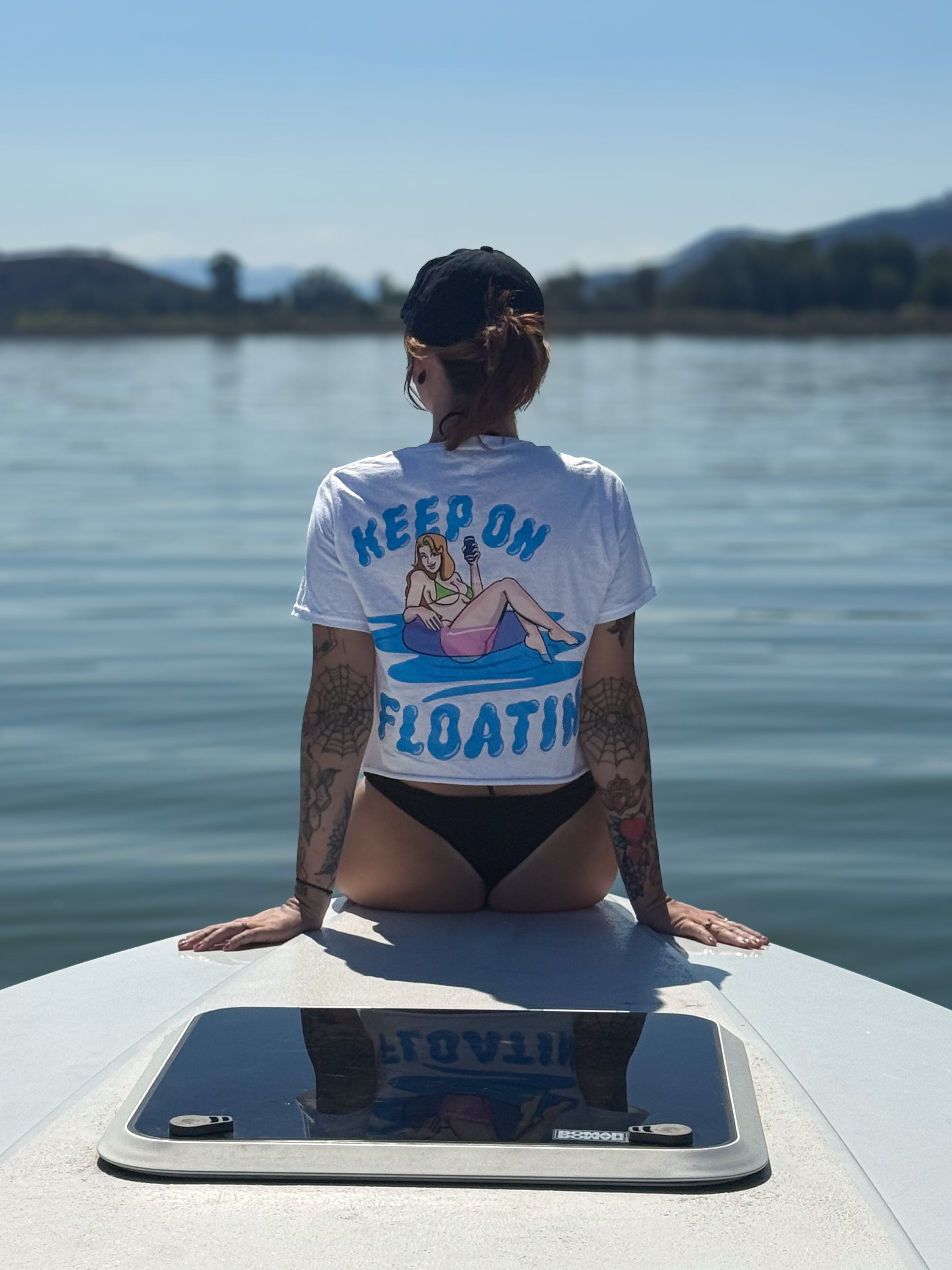 Person sitting on a boat wearing a t-shirt with a graphic design and text, looking out over water.